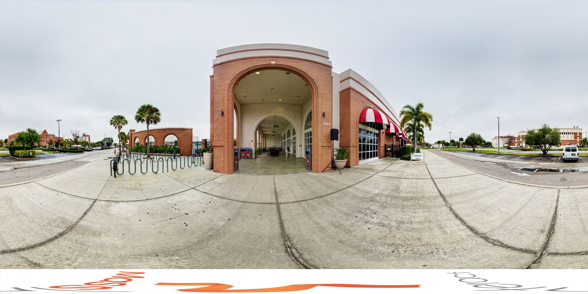 Panoramic view of a college campus featuring Panther Dining Hall with a large arched entrance, bicycle racks, and surrounding palm trees on an overcast day